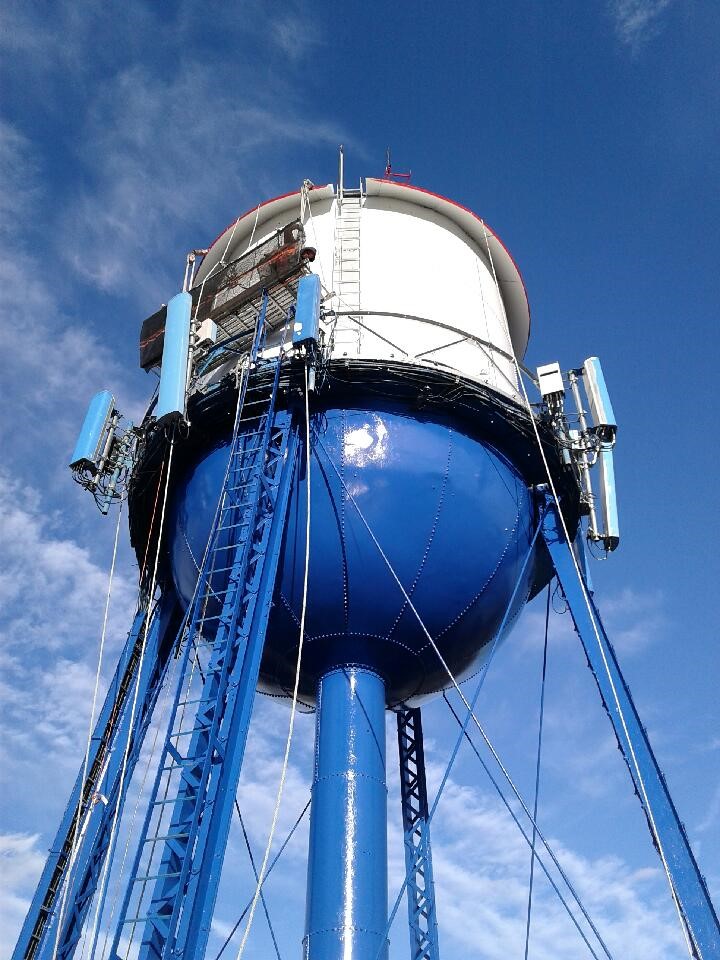 Blue and white-water tower with cellular antennas, ladder access, and cable support structure against a clear sky.