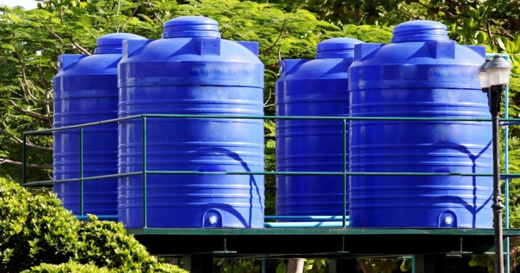 Four blue plastic water storage tanks on metal framework platform surrounded by lush green vegetation.