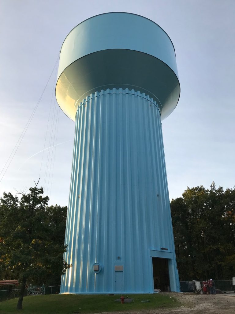 Tall light blue cylindrical water tower with mushroom-shaped top and ribbed column design against tree line.